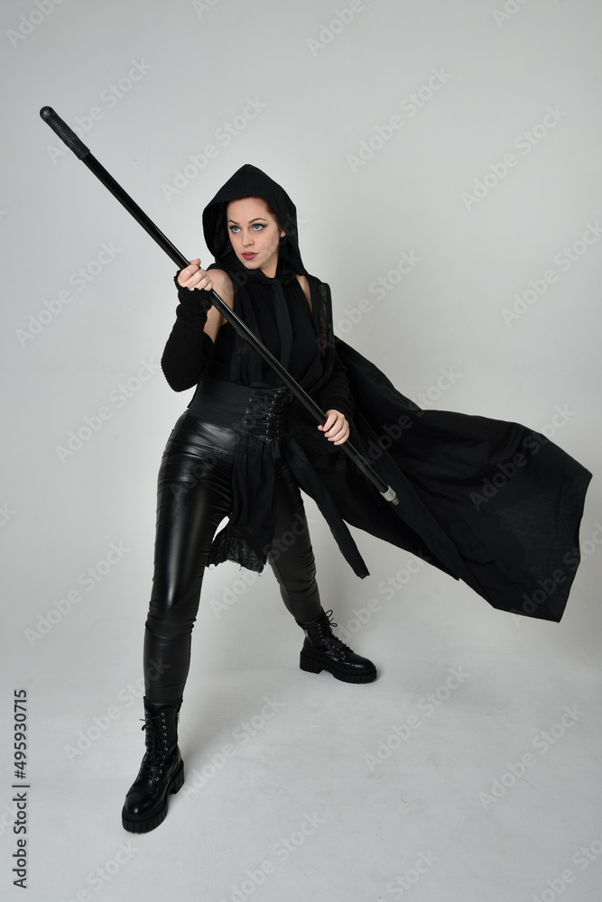 Full length portrait of pretty redhead female model wearing black futuristic scifi leather costume, black cloak holding a spear weapon. Dynamic standing pose on white studio background. Full length portrait of pretty redhead female model wearing black futuristic scifi leather costume, black cloak holding a spear weapon. Dynamic standing pose on white studio background.