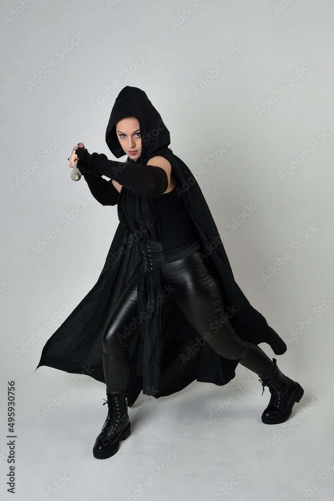 Full length portrait of pretty redhead female model wearing black futuristic scifi leather costume, black cloak holding a spear weapon. Dynamic standing pose on white studio background.