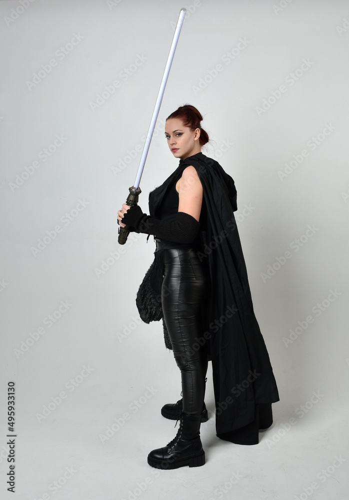 Full length portrait of pretty redhead female model wearing black futuristic scifi leather cloak costume, holding a gun weapon. Dynamic standing pose on studio background. Full length portrait of pretty redhead female model wearing black futuristic scifi leather cloak costume, holding a gun weapon. Dynamic standing pose on studio background.