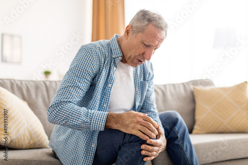 Mature man with knee pain sitting on couch at home photo