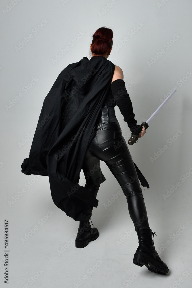 Full length portrait of pretty redhead female model wearing black futuristic scifi leather cloak costume, holding a lightsaber weapon. Dynamic standing pose on white studio background. Full length portrait of pretty redhead female model wearing black futuristic scifi leather cloak costume, holding a lightsaber weapon. Dynamic standing pose on white studio background.