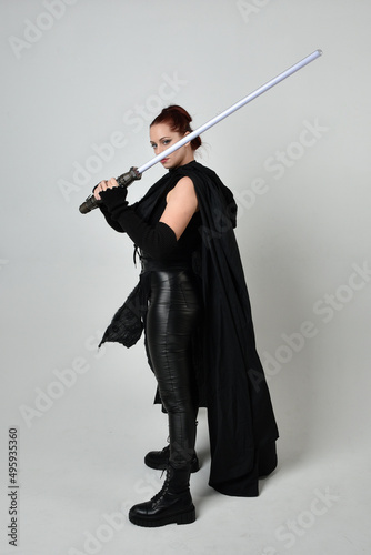 Full length portrait of pretty redhead female model wearing black futuristic scifi leather cloak costume, holding a lightsaber weapon. Dynamic standing pose on white studio background.