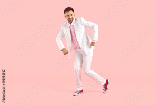 Cheerful trendy guy in white formal suit funny dancing isolated on pastel pink background. Full length of funny man in suit and sneakers. Concept of leisure, weekend, cheerful mood, joy and happiness.