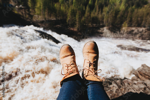 Woman's legs in blue jeans and light brown boots sitting on the edge near waterfall