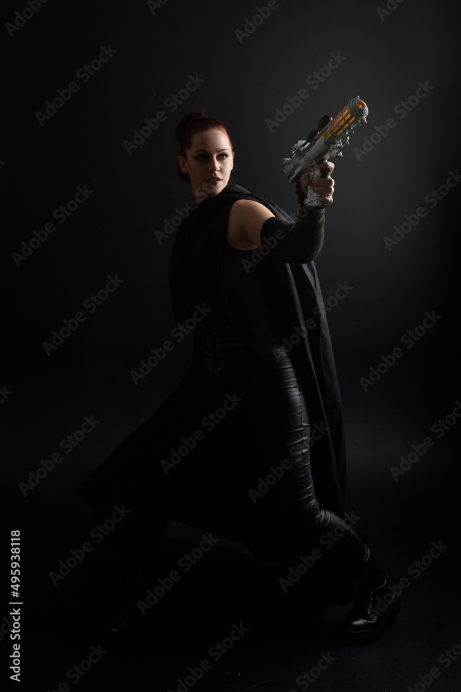 Fototapeta premium Full length portrait of pretty redhead female model wearing black futuristic scifi leather cloak costume, holding a gun weapon. Dynamic standing pose on studio background.