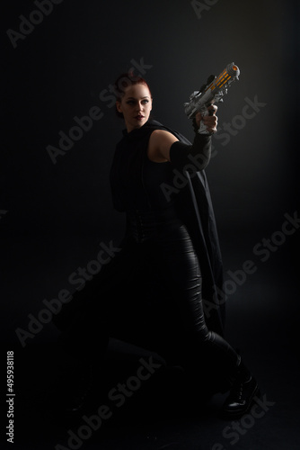 Full length portrait of pretty redhead female model wearing black futuristic scifi leather cloak costume, holding a gun weapon. Dynamic standing pose on studio background.