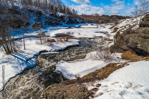 A spring landscape with a river, rocks, ice, snow, trees, a ruined brick building and a blue sky with white clouds
