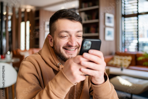 Cheerful freelancer with beard browsing smartphone and smiling while sitting at table with mobile phone in light cafe in daytime.