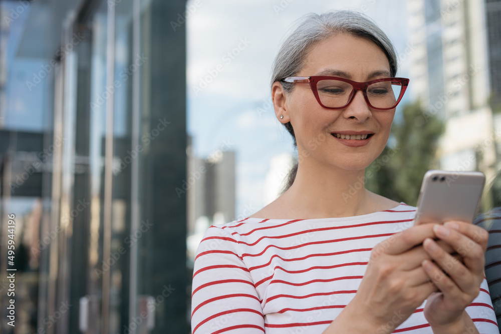Smiling mature businesswoman using mobile phone communication online, searching on website, reading email on the street. Confident asian female wearing red stylish eyeglasses shopping online outdoors 