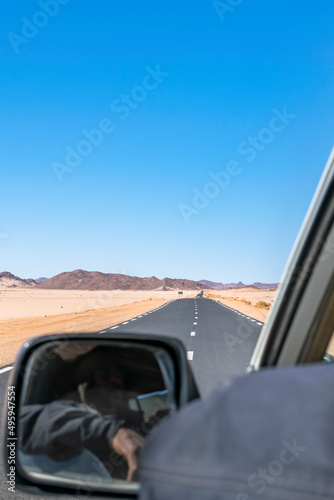 Long Sahara road trip in empty reg and sandy desert, rocky mountains seen from the rear side a moving car, tuareg driver non recognisable is visible in the side mirror his arm outside the 4x4 vehicle.