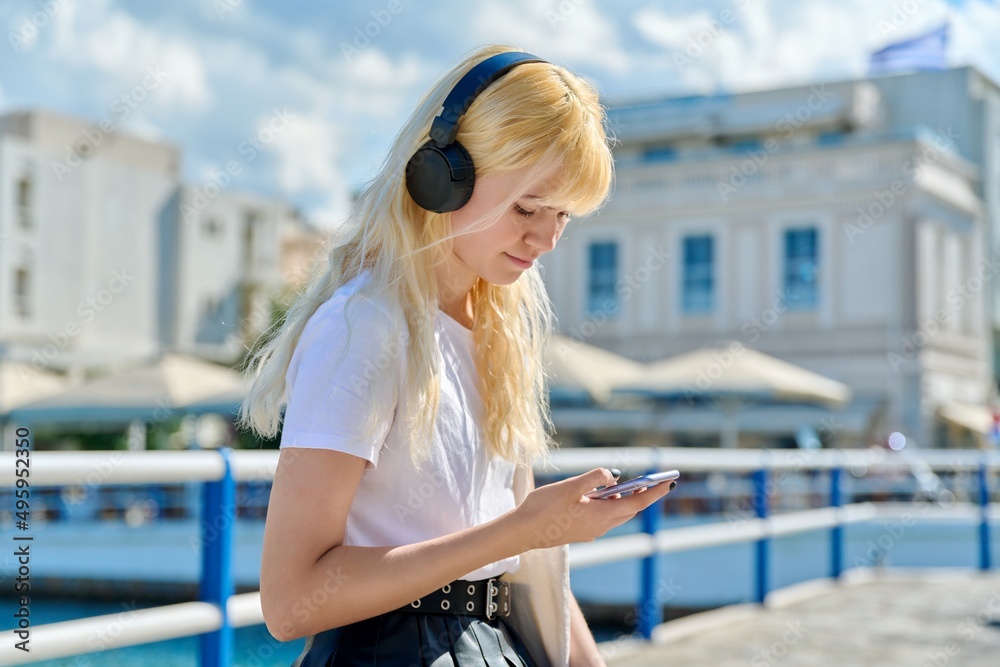 Beautiful teenage female in headphones reading looking at smartphone screen outdoor