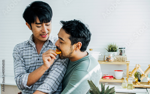 Gay LGBT sweet Asian couple wearing pajamas, smiling with happiness and love, eating, feeding orange, healthy fruits for breakfast in kitchen at home in the morning with sunlight. Lifestyle Concept.
