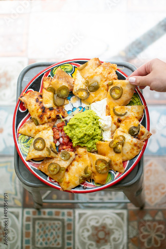 Hand grabs a nacho from a plate full of nachos with melted cheese, jalapenos, gaucamole, pico de gallo and guacamole. Colorful plate. Zenital. Sunlight. photo