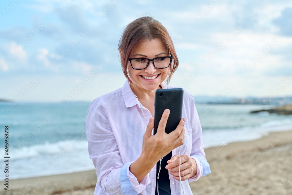 Happy middle aged woman on sea vacation enjoying in headphones with smartphone