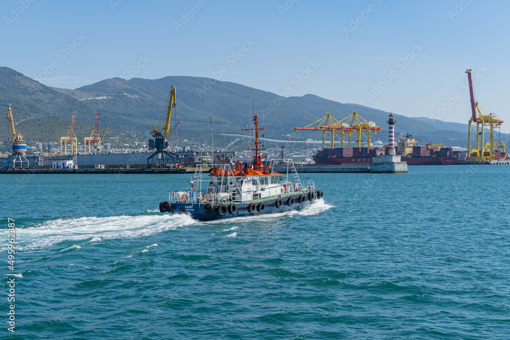 Blue pilot tug is moving along bay. In background are the piers, port cranes and mountains of Tsemess Bay. Close-up. Novorossiysk commercial sea port. Novorossiysk, Russia - September 15, 2021