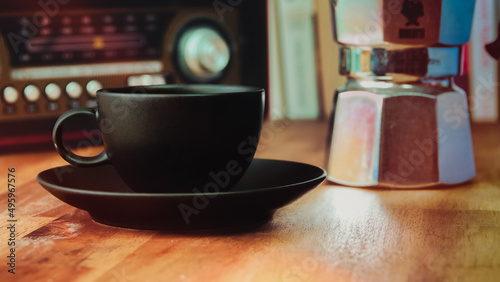 Moka pot, vintage radio, a black coffee cup and saucer on a brown wooden table, with books in the background, vintage look 