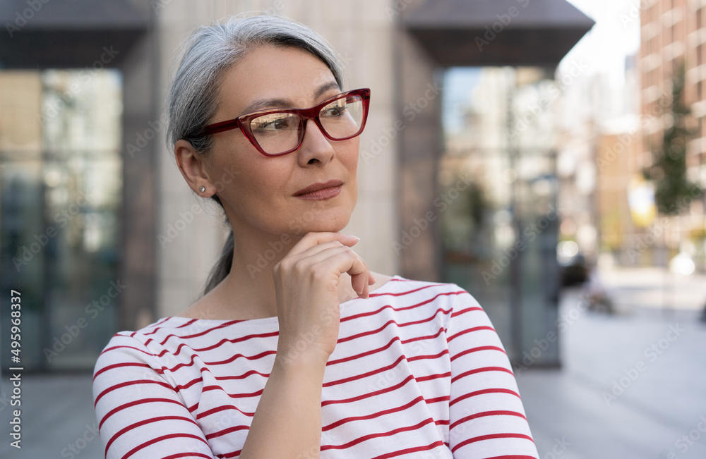 Confident mature businesswoman wearing red stylish eyeglasses standing on urban street, copy space. Portrait of pensive asian manager waiting for taxi looking away outdoors. Successful business 