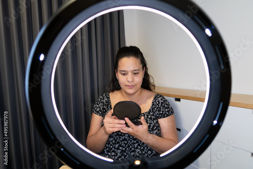 joven mujer se mira al espejo frente a aro de luz en interior de su recámara photo