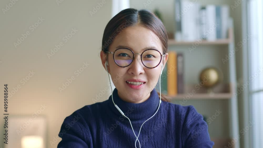 Young Asian woman creator wearing glasses and headphone talking on video call conference or virtual meeting at home, look at camara webcam view