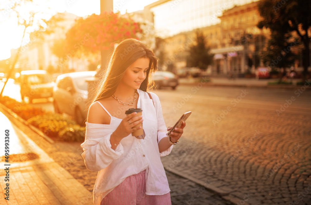 Fototapeta premium long-haired girl in glasses with phone, coffee and bag walks around the city at sunset