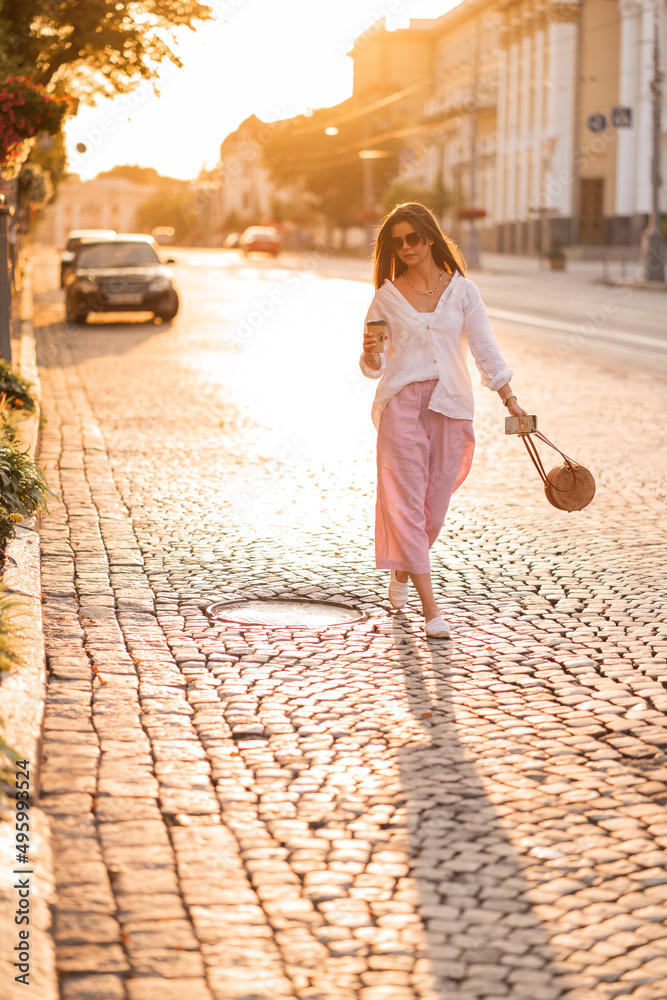 long-haired girl in glasses with phone, coffee and bag walks around the city at sunset