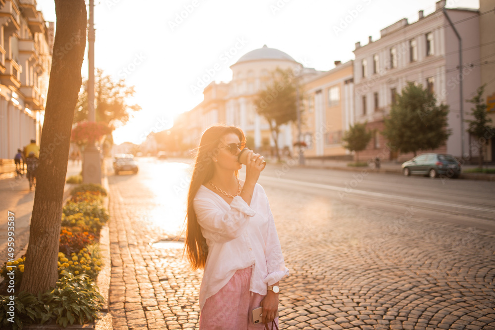 long-haired girl in glasses with phone, coffee and bag walks around the city at sunset