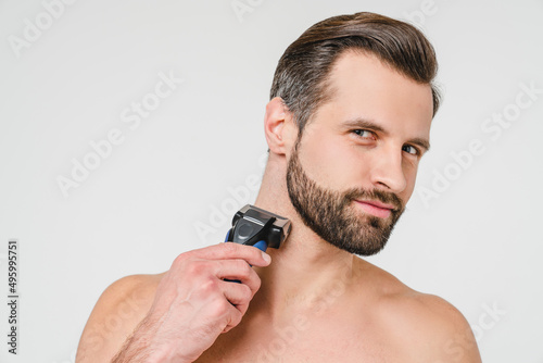 Caucasian attractive young man shaving his beard with electric razor, doing barber shop face care, doing morning hygiene preparations for male beauty isolated in white background