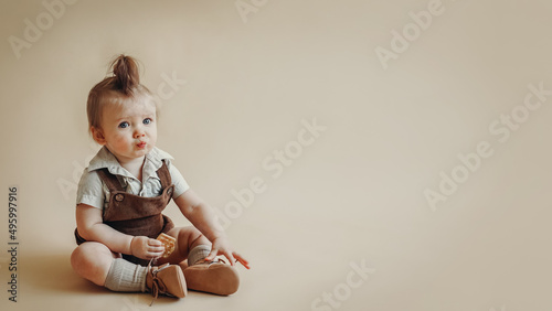 baby in a brown vintage corduroy jumpsuit, shorts, knee socks sits on a beige background and eats cookies, stretching his lips funny