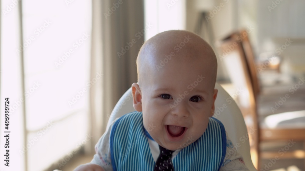 Childhood, Happiness, family, Motherhood. Cheerful happy smiling baby Close up face. Emotions of newborn boy. child in a bib sits in a feeding chair gnaws toothpick in the kitchen, Infant gum pain