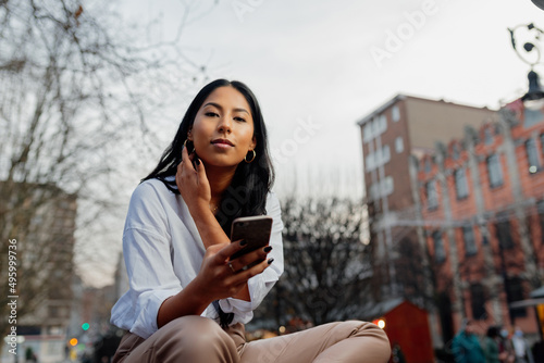 portrait of young latin woman looking at camera and holding her mobile phone in a city street.