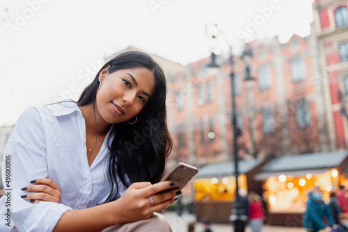 Canvas Print portrait of young latin woman looking at camera and holding her mobile phone in a city street