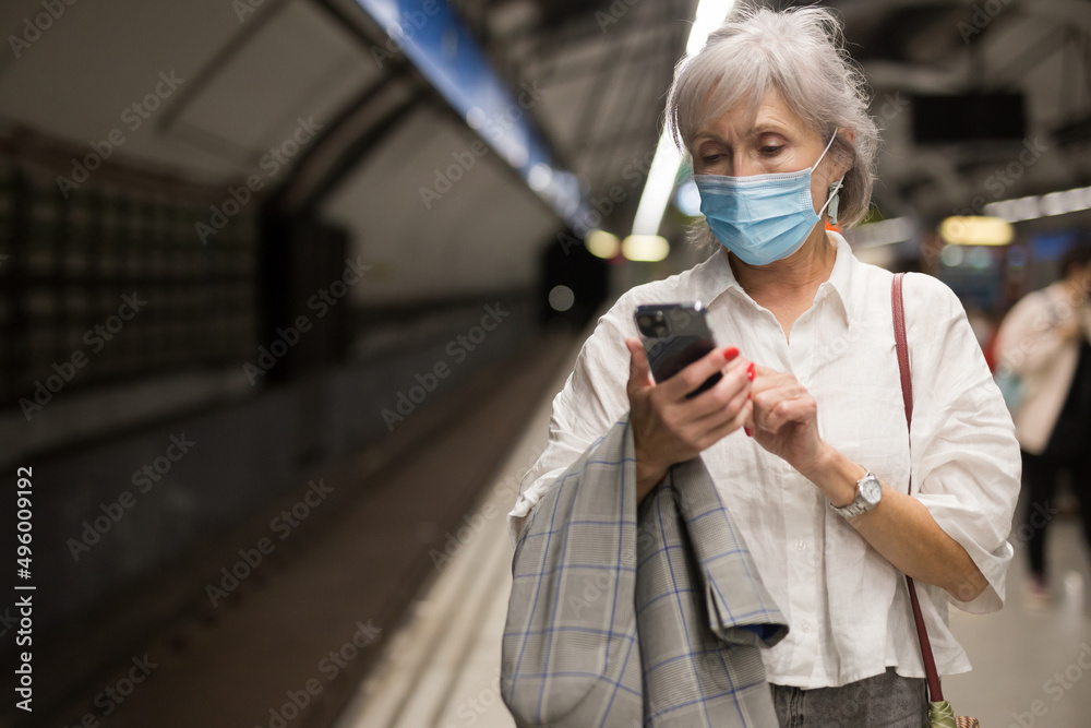 Senior woman in face mask with cell phone in hand standing in subway station near railway and waiting for train.