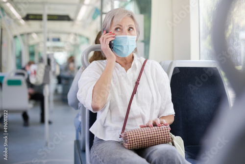Gray haired caucasian woman in face mask sitting in tram and having telephone conversation.