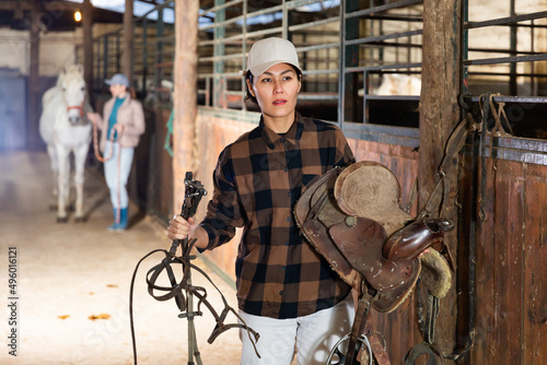 Asian woman leads white horse out of the stall. Another woman carries saddle for a horse