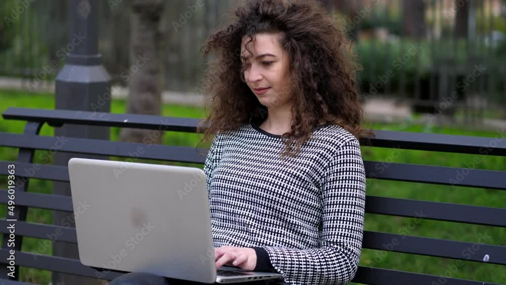 Young curly woman working in the park during coronavirus social isolation. Smiling young woman using laptop sitting on the bench in park outdoors Young curly woman working in the park during coronavirus social isolation. Smiling young woman using laptop sitting on the bench in park outdoors