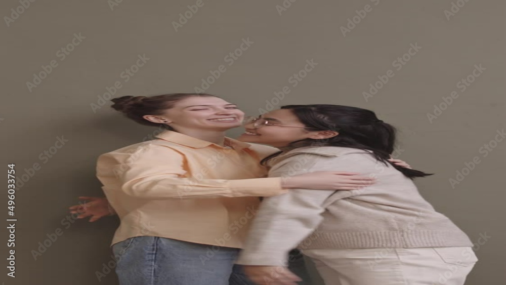 Vertical medium slowmo portrait of two young cheerful multiethnic girlfriends hugging each other and smiling at camera standing on pastel olive green background Vertical medium slowmo portrait of two young cheerful multiethnic girlfriends hugging each other and smiling at camera standing on pastel olive green background