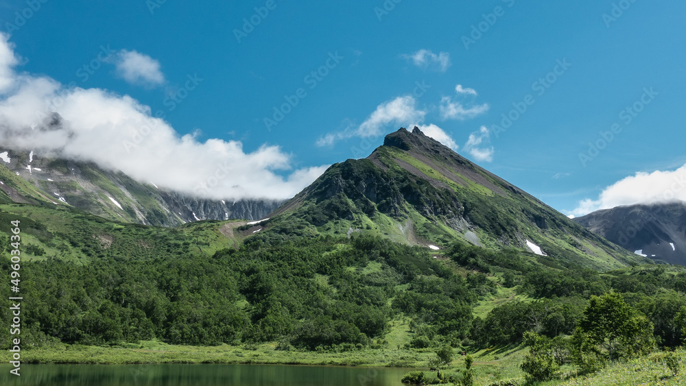 The picturesque mountain range is covered with green vegetation. Patches of melted snow are visible on the slopes. There is an emerald lake in the valley. Clouds in the blue sky. Kamchatka.
