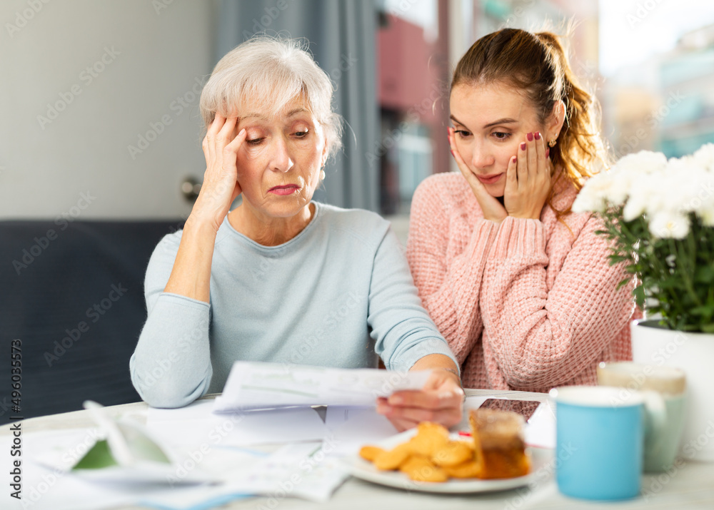Worried elderly woman sitting in dining room with adult daughter, checking utility bills.