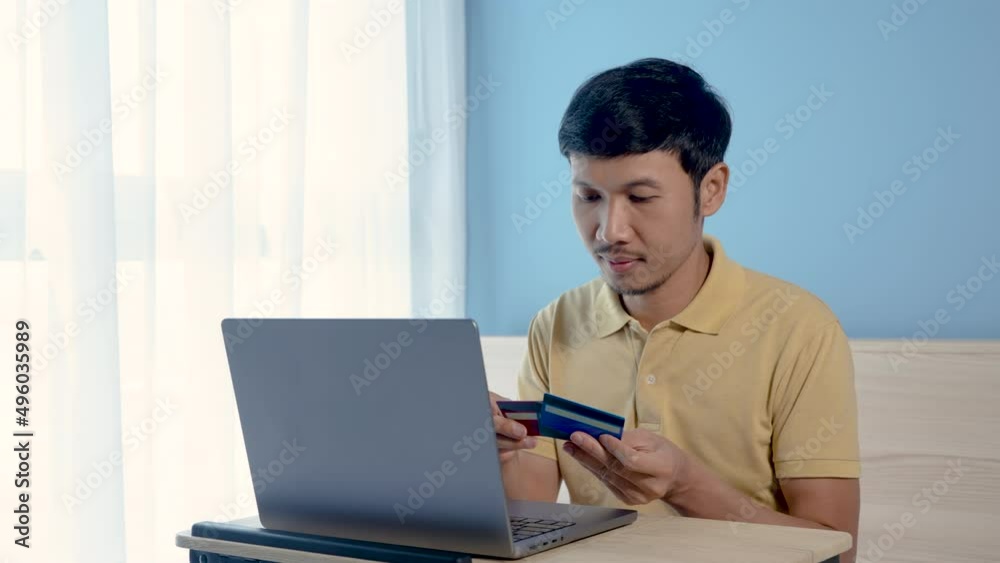 Handsome Asian man, wearing a yellow shirt sitting on a white bed, has a serious look on his face when he chooses a credit card to pay for an order online, on his laptop computer. Handsome Asian man, wearing a yellow shirt sitting on a white bed, has a serious look on his face when he chooses a credit card to pay for an order online, on his laptop computer.