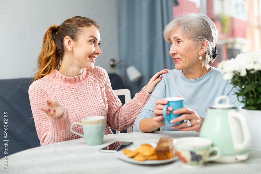 Friendly family of elderly mother and young adult daughter having good time while drinking tea and chatting happily in living room.