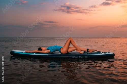 A young slim sexy tanned woman in swimsuit lies on a sup board. Sunset in the background. Copy space. Concept of summer water sport