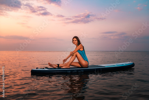Pretty caucasian woman in swimsuit sitting on a sup board. Sunset in the background. Copy space. Concept of water sport and summer activity © _KUBE_