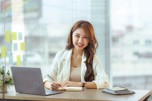 Charming Asian woman with a smile sitting and taking notes with computer laptop on her desk, enjoying work. Looking at camera.