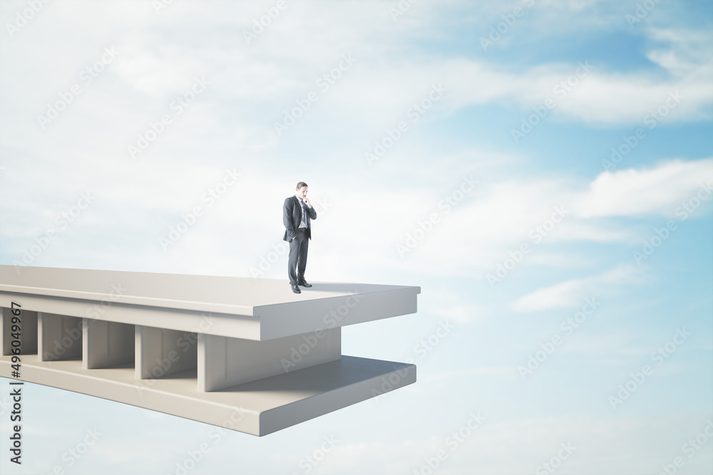 Attractive pensive young man standing on bridge with gap and blue sky background with mock up place. Challenge and obstacle concept. Attractive pensive young man standing on bridge with gap and blue sky background with mock up place. Challenge and obstacle concept.