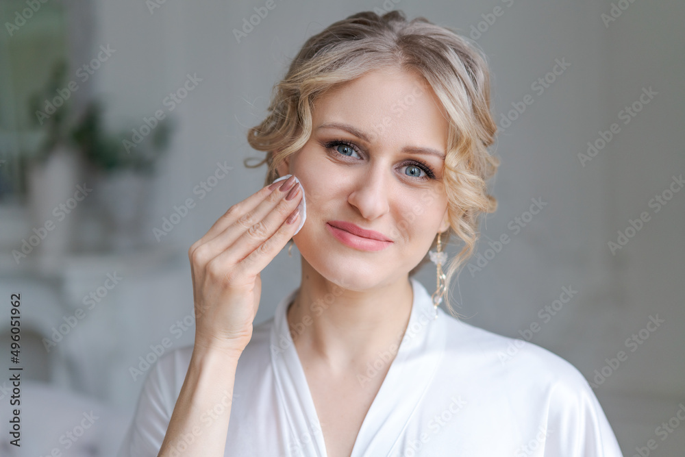 Skin care, make-up removal, cleansing and routine at home. Smiling caucasian woman in a white bathrobe wipes her face with a cotton sponge in the interior of the bedroom, empty space Skin care, make-up removal, cleansing and routine at home. Smiling caucasian woman in a white bathrobe wipes her face with a cotton sponge in the interior of the bedroom, empty space