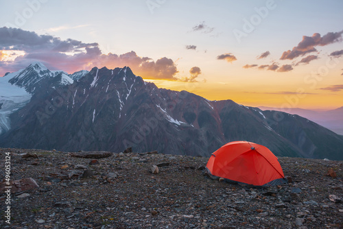 Scenic alpine landscape with tent at very high altitude with view to large mountains in orange dawn sky. Vivid orange tent with awesome view to high mountain range under cloudy sky in sunset colors.