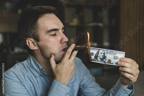 Young brutal businessman lighting cigar with 100 dollar bill as symbol of wealth and success. concept of wealth and extravagance. photo