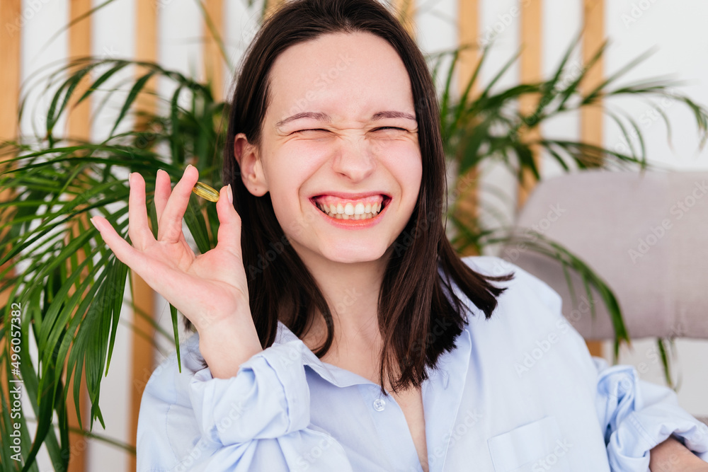 Smiling young woman with short hair holding pill omega capsule in hand at home. Girl taking medicine indoors Smiling young woman with short hair holding pill omega capsule in hand at home. Girl taking medicine indoors
