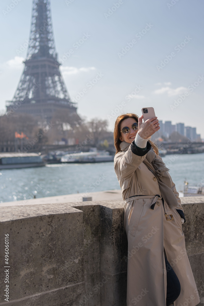Happy young woman traveler makes selfie portrait on mobile phone against Eiffel tower in Paris. Architecture, attraction, landmark. Vacation, travelling, trip.