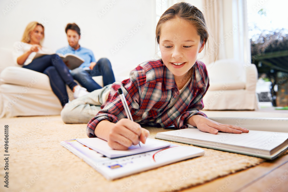 Homework done equals relaxing time. Shot of a little girl lying on the living room floor doing homework with her parents in the background. Homework done equals relaxing time. Shot of a little girl lying on the living room floor doing homework with her parents in the background.